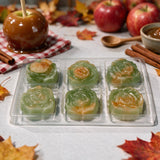 Six green rose-shaped jelly candies in a clear plastic tray, styled with caramel apples, red apples, cinnamon sticks, and autumn leaves on a white tablecloth.
