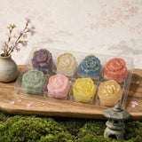 A clear plastic tray holding eight rose-shaped wax melts or soaps in assorted pastel and metallic colors, arranged on a wooden board with moss, a small vase of flowers, and a stone lantern against a soft floral background.