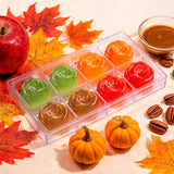 A clear plastic tray holding eight rose-shaped gel desserts in assorted colors, styled with autumn leaves, pumpkins, coffee beans, a red apple, and a small bowl of caramel on a light surface.